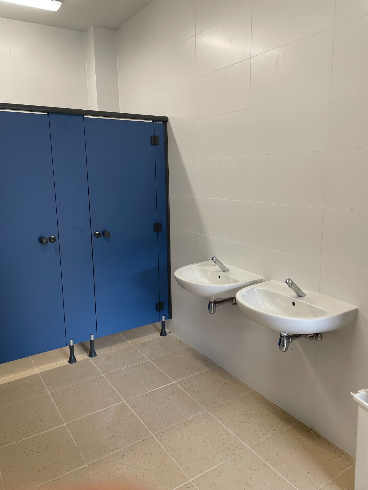 Gerenoveerde sanitaire schoolruimte in Gent met blauwe cabines, lichte tegelvloer en twee wastafels.