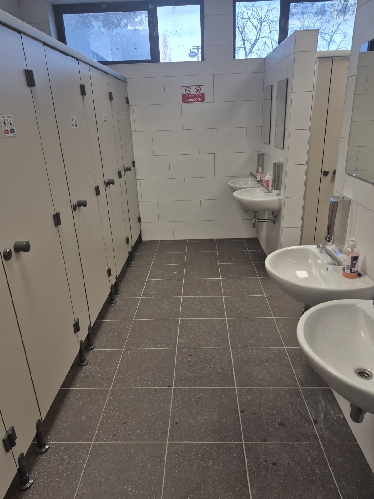 Gerenoveerde sanitaire schoolruimte in Brussel met grijze vloertegels, witte wandtegels, cabines en wastafels.