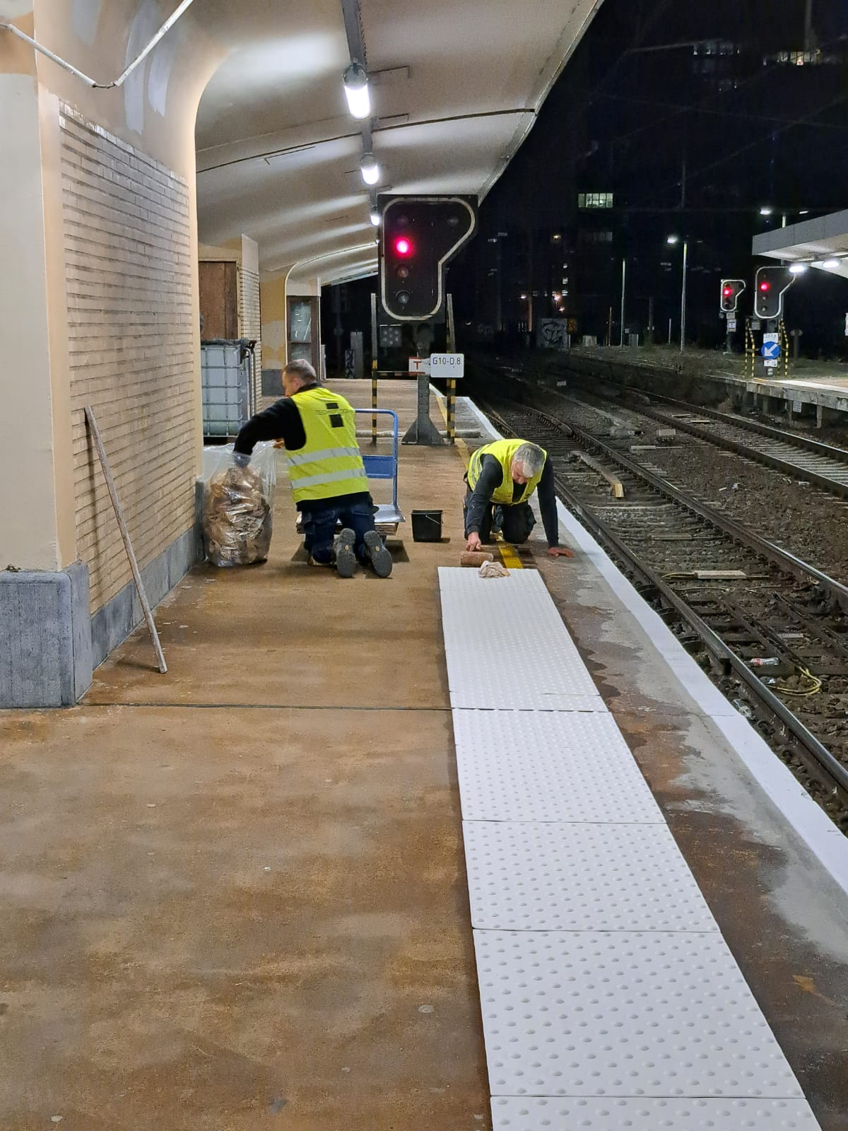 Plaatsing van kunststof kleeftegels op een treinperron in Brussel-Noord.